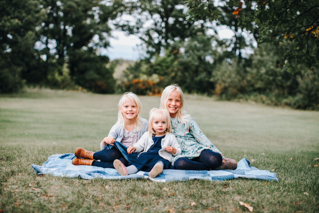 three blond girls sitting on a denim blanket on the grass